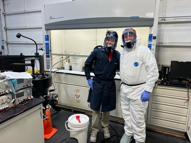 2 students in full PPE on in a white suit and gas mask and the other in a black suit and gas mask